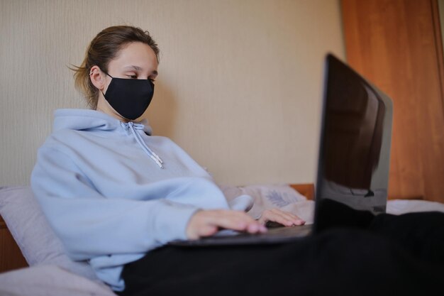 Kaukasisches Mädchen in blauem Hoodie und schwarzer Maske arbeitet isoliert am Laptop zu Hause