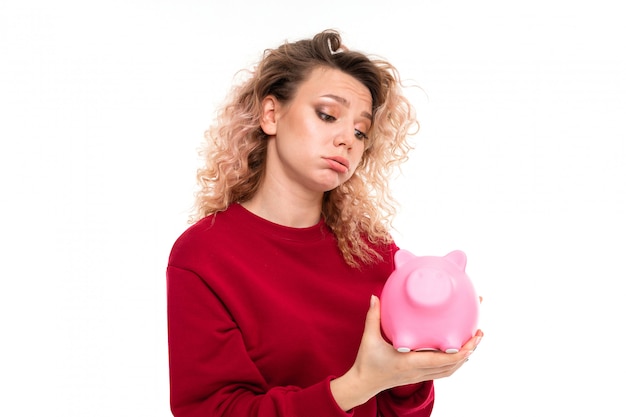 Kaukasisches Mädchen mit dem lockigen angemessenen Haar hält ein rosafarbenes Schwein moneybox, das Portrait an, das auf Weiß getrennt wird