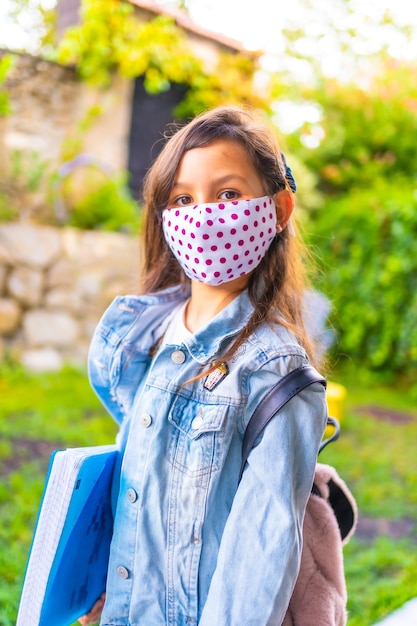 Kaukasisches Mädchen mit Gesichtsmaske bereit, zur Schule zurück zu gehen. Neue Normalität, soziale Distanz, Coronavirus-Pandemie, Covid-19. Jacke, Rucksack, Maske mit rosa Punkten und einem blauen Block in der Hand