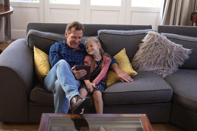 Kaukasisches Mädchen und ihr Großvater genießen die gemeinsame Zeit in einem Haus, sitzen auf einer Couch, umarmen sich und nutzen ein digitales Tablet