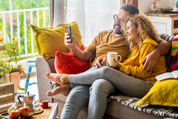 Foto kaukasisches paar, das zu hause einen videoanruf mit dem handy macht, der auf dem sofa sitzt mann und frau, die beim morgendlichen frühstück eine drahtlose technologieverbindung genießen und spaß daran haben, freunde anzurufen