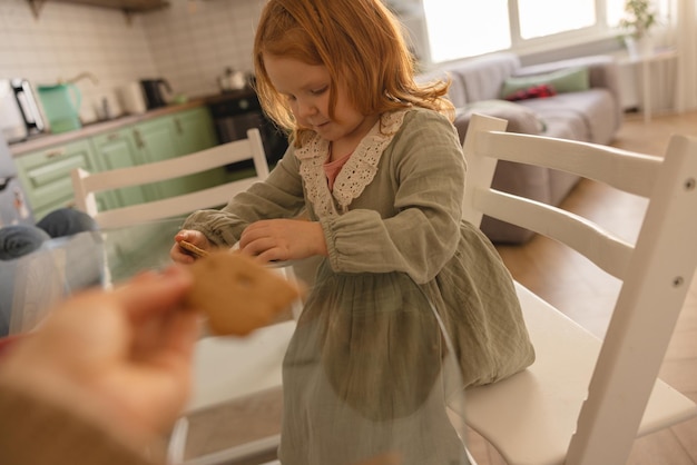 Foto kaukasisches rothaariges kleinkind kaut kekse mit vergnügen und vergnügen, während es am tisch sitzt snack-konzept