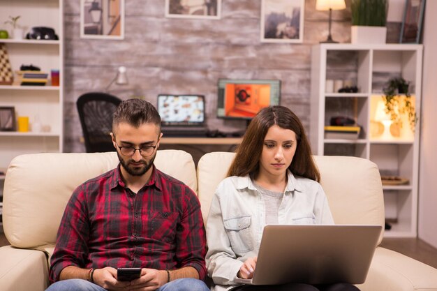 Kaukasisches schönes Paar mit ernsten Gesichtern, die auf Sofa sitzen. Paar mit Laptop und Telefon beim Sitzen auf dem Sofa.