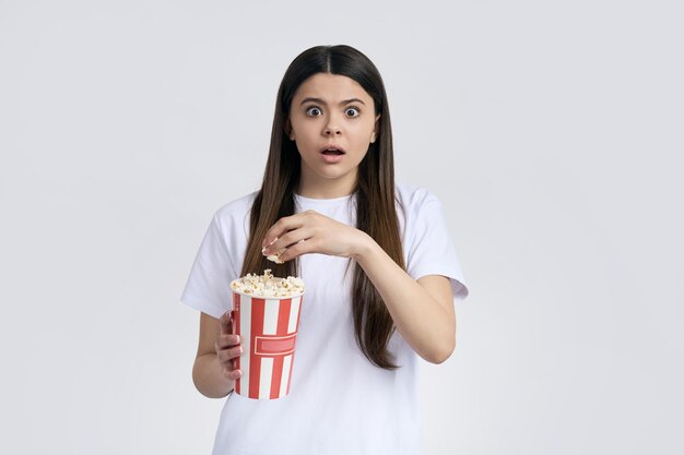 Kaukasisches süßes Teenager-Mädchen in weißem T-Shirt-Attrappe, das einen Pappeimer mit leckerem Popcorn in der Hand hält und ängstlich und ängstlich aussieht und sich einen Gruselfilm vor isoliertem weißem Hintergrund ansieht. Menschen und Kino