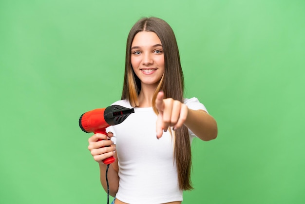 Kaukasisches Teenagermädchen, das einen Haartrockner über isoliertem Hintergrund hält, zeigt mit einem selbstbewussten Ausdruck mit dem Finger auf Sie
