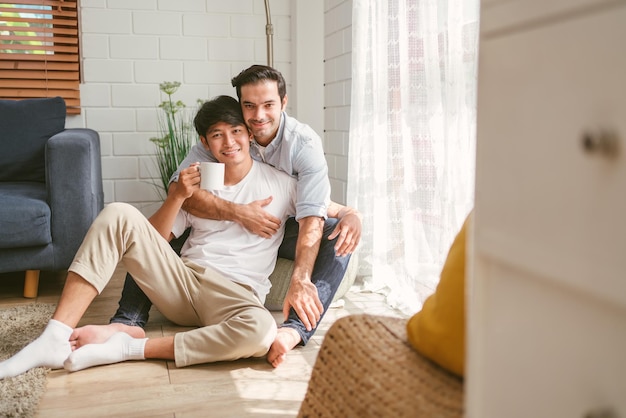Kaukasisches und asiatisches LGBT-Paar, das sich von hinten umarmt, sitzt zu Hause zusammen und trinkt eine Tasse Kaffee. Vielfalt der LGBT-Beziehungen. Ein Konzept für das Zusammenleben eines schwulen Paares