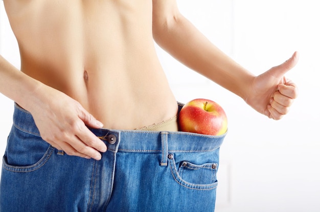 Kaukasisches weibliches Modell in Blue Jeans mit rotem Apfel, der ihren flachen Bauch zeigt. Gesunder Lebensstil und Weightloss-Konzept.