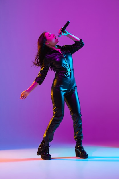 Kaukasisches weibliches Sängerporträt lokalisiert auf purpurrotem Studiohintergrund im Neonlicht. Schönes weibliches Modell in schwarzer Kleidung mit Mikrofon. Konzept der menschlichen Emotionen, Gesichtsausdruck, Anzeige, Musik, Kunst.