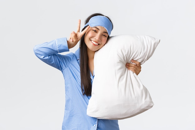 Kawaii glücklich lächelndes asiatisches Mädchen in blauer Schlafmaske und Pyjama, Kissen umarmt und Friedenszeichen zeigend, sich nach guter Nachtruhe wohlfühlen, während der Übernachtung im Bett liegend, weißer Hintergrund.