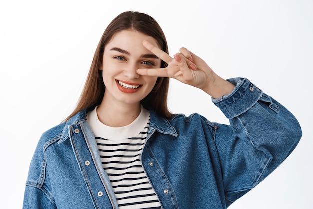 Kawaii positives Mädchen zeigt Peace Vsign in der Nähe des Auges, lächelt und sieht glücklich aus, wenn es in Freizeitkleidung vor weißem Hintergrund steht