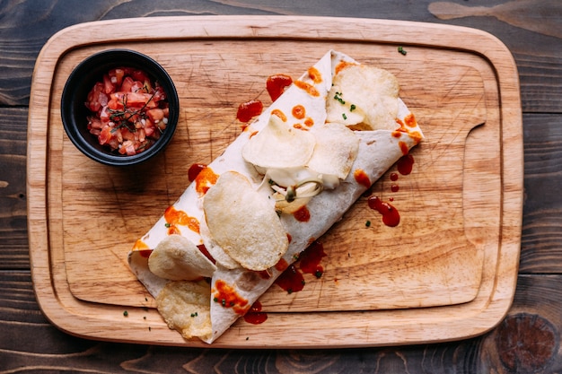 Kebab de carne con patatas fritas relleno con salsa y servido con ensalada de tomate en placa de madera.