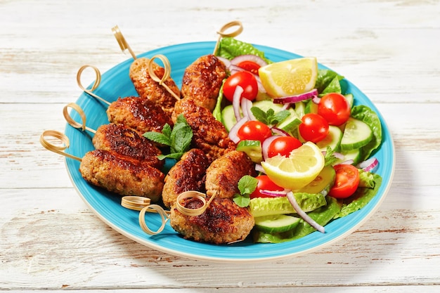 Kebabs de cordero kofta de cordero molido, servidos en un plato azul con aderezo de yogur, ensalada de verduras frescas y gajos de limón, cocina griega en una mesa de madera blanca, vista superior, primer plano