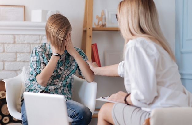 Keine Sorgen. Verärgerter Junge mit modischer Frisur, der kariertes Hemd und Jeans trägt und seine Hände auf seinen Augen hält, während er auf weißem Stuhl sitzt