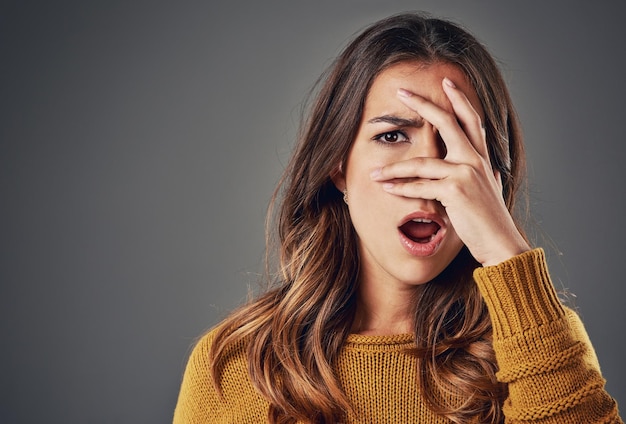 Keine Worte können ihre Gefühle im Moment ausdrücken Porträt einer jungen Frau, die vor einem grauen Hintergrund durch ihre Finger späht