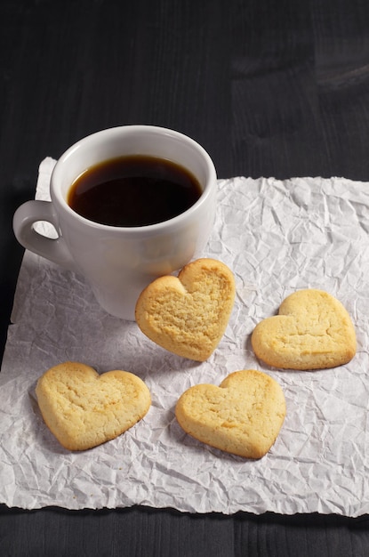 Kekse in Form eines Herzens und einer Tasse Kaffee auf einem zerknitterten Papier befinden sich auf einem schwarzen Holzhintergrund