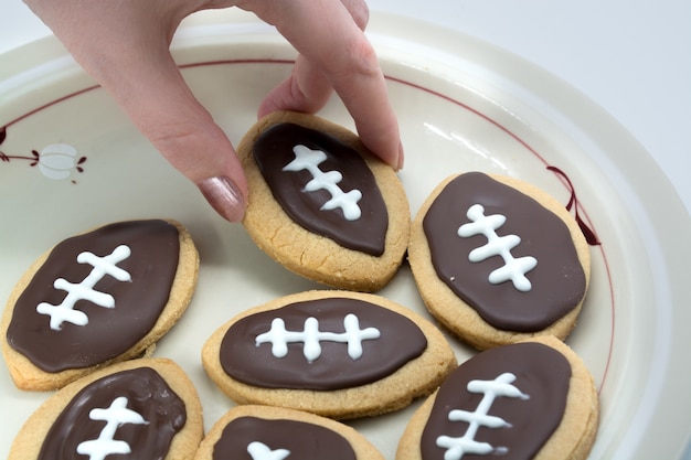 Kekse in Fußballform. Hand, die Plätzchen vom Teller nimmt. Amerikanisches Sportkonzept. Super Bowl-Partykekse. Foto in hoher Qualität