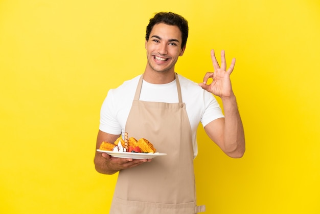 Kellner im Restaurant, der Waffeln über isoliertem gelbem Hintergrund hält, der ein OK-Zeichen mit den Fingern zeigt