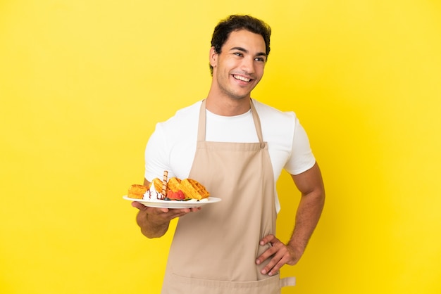 Kellner im Restaurant, der Waffeln über isoliertem gelbem Hintergrund hält, posiert mit den Armen an der Hüfte und lächelt