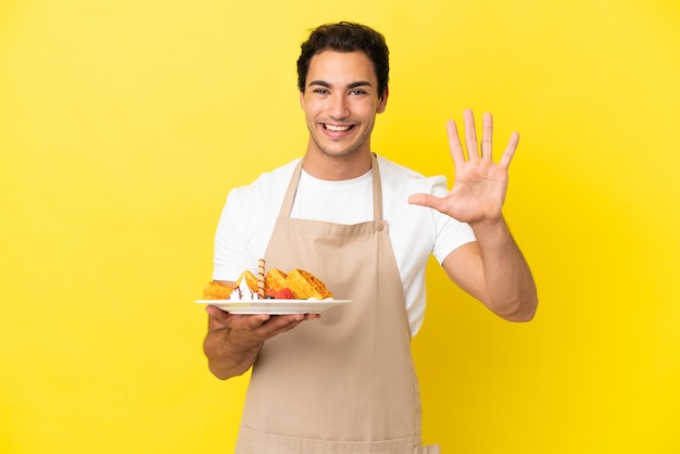 Kellner im Restaurant hält Waffeln über isoliertem gelbem Hintergrund und zählt fünf mit den Fingern