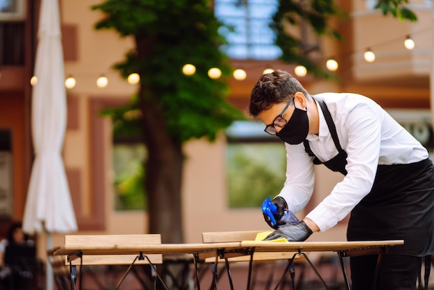 Kellner in schützender Gesichtsmaske und Handschuhen, die den Restauranttisch für den nächsten Kunden reinigen