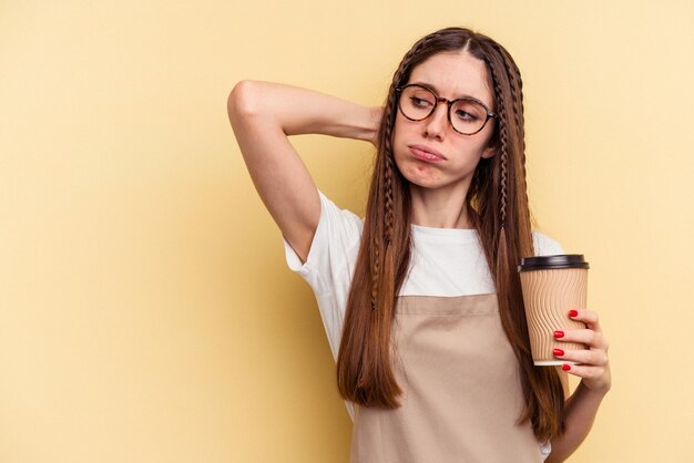 Kellnerin im Restaurant, die einen Kaffee zum Mitnehmen hält, isoliert auf gelbem Hintergrund, der den Hinterkopf berührt und nachdenkt und eine Wahl trifft