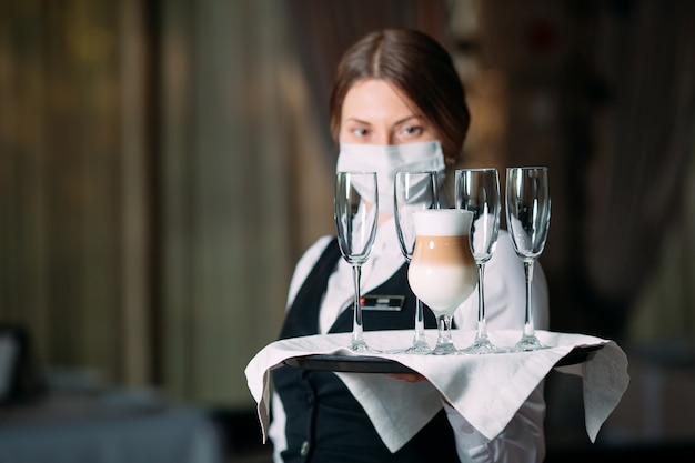 Foto kellnerin mit europäischem aussehen in einer medizinischen maske serviert latte-kaffee.