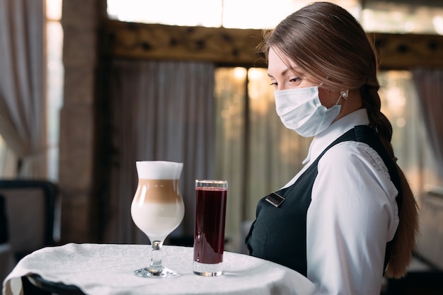 Kellnerin mit europäischem Aussehen in einer medizinischen Maske serviert Latte-Kaffee.