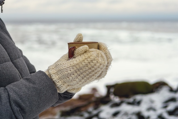 Keramikbecher in den Händen einer Frau in weißen Strickhandschuhen auf einer verschwommenen Schneelandschaft.