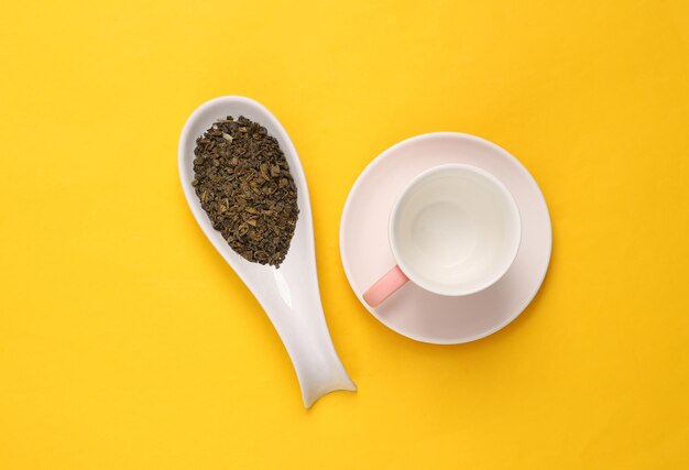 Keramischer Löffel mit trockenem grünem Tee und Tasse auf gelben Hintergrund