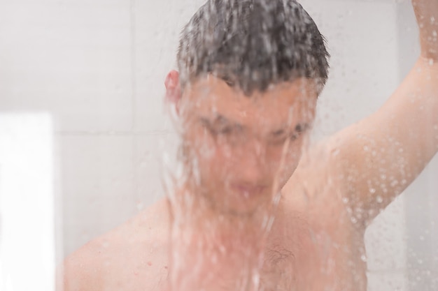 Kerl, der duscht und den Duschkopf hält, während er unter fließendem Wasser hinter einer durchsichtigen beschlagenen Glastür im Badezimmer steht