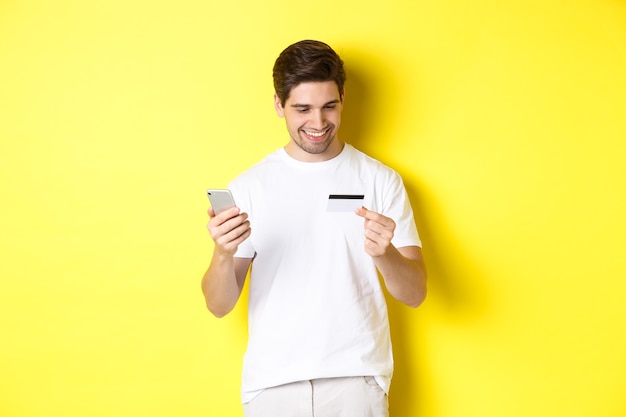 Kerl, der Online-Bestellung macht, registriert Kreditkarte in der mobilen Anwendung, hält Smartphone und lächelt, steht über gelber Wand