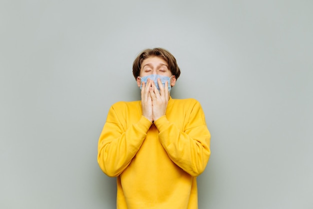 Kerl in einem gelben Sweatshirt und einer medizinischen Maske niest auf einem grauen Hintergrund mit geschlossenen Augen