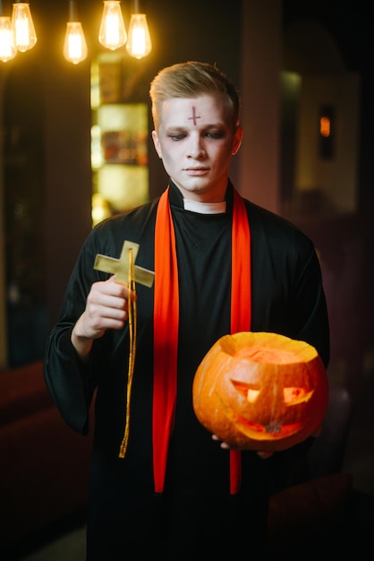 Kerl in einem Halloween-Priesterkostüm mit ernstem Gesicht hält einen geschnitzten Kürbis in der Hand Die andere Hand hebt ein religiöses Kreuz Ein junger Mann schaut in die Kamera