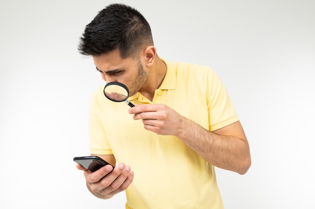 Kerl in einem T-Shirt schaut mit einer Lupe das Telefon in seiner Hand auf einem weißen Hintergrund aus.