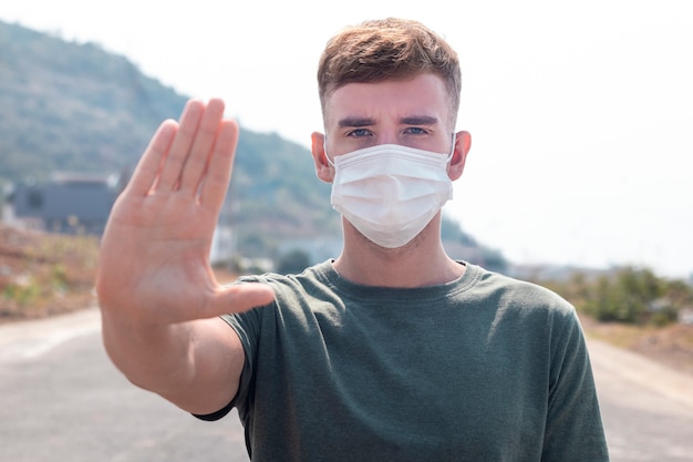 Foto kerl, junger mann in der schützenden sterilen medizinischen maske auf seinem gesicht betrachten kamera draußen, auf asiatischer straßenshowpalme, hand, stoppen sie kein zeichen. luftverschmutzung, virus, pandemie coronavirus-konzept. covid-19