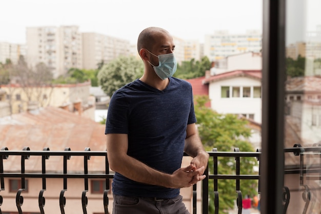 Kerl mit Gesichtsmaske, der sich während der Selbstisolation auf dem Balkon der Wohnung entspannt.