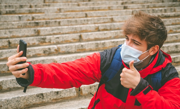 Kerl mit Gesichtsmaske und Rucksack, der an einem Wintertag ein Selfie auf der Straßentreppe macht