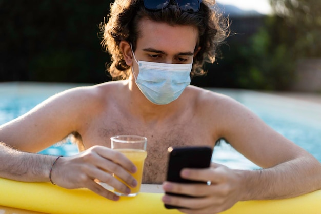 Kerl mit medizinischer Gesichtsmaske, der sich am Pool amüsiert, mit dem Smartphone tippt und im Sommer 2021 nach dem Ausbruch des Coronavirus Orangensaft trinkt Kerl genießt das Leben allein aufgrund von Sommerbeschränkungen