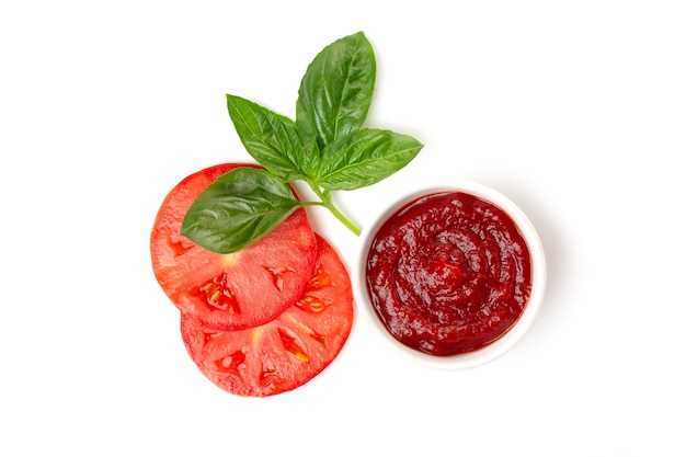 Ketchup (pasta de tomate) en una cacerola con hojas de albahaca y rodajas de tomate sobre un fondo blanco.