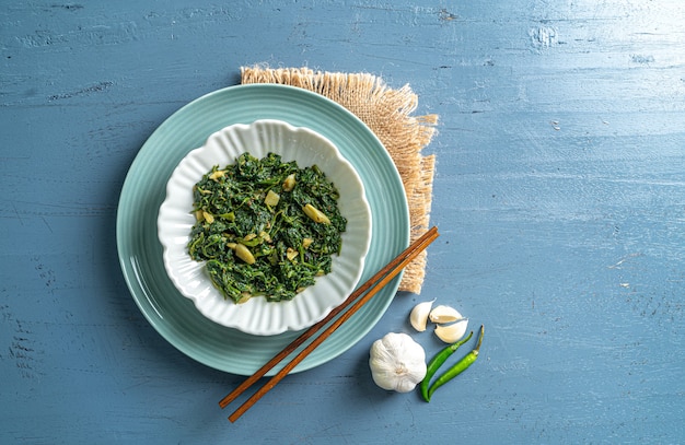 Keto-Diät-Spinat-Curry in weißer Schüssel und blauer Platte mit Essstäbchen und Knoblauch-Chilis mit Vintage-Schicht auf blauer Holztisch-Draufsicht