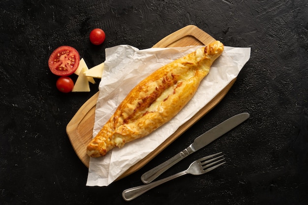 Khachapuri com queijo e tomate em uma placa de corte de madeira ao lado de talheres e ingredientes em uma mesa preta. vista do topo.