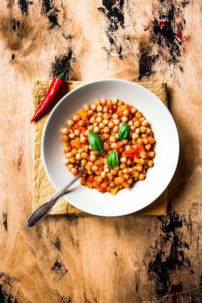 Kichererbseneintopf mit Tomaten, gelbem Pfeffer und Chili, Basilikumblättern. Draufsichtfoto, Holzoberfläche. Löffel in einen Teller.