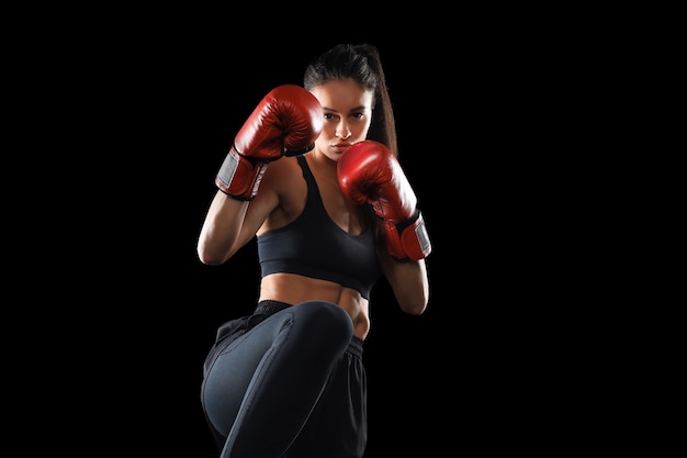 Kickbox-Frau in Activewear und roten Kickbox-Handschuhen auf schwarzem Hintergrund, die einen Kampfsport-Kick ausführt. Sportübungen, Fitnesstraining.