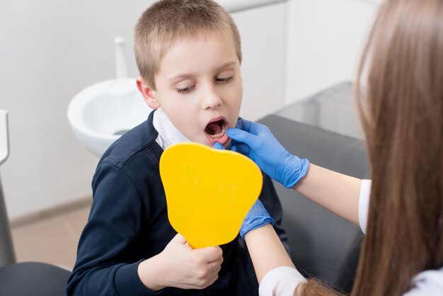 Kid boy en la silla del dentista se mira en el espejo y el médico con guantes azules le muestra los dientes