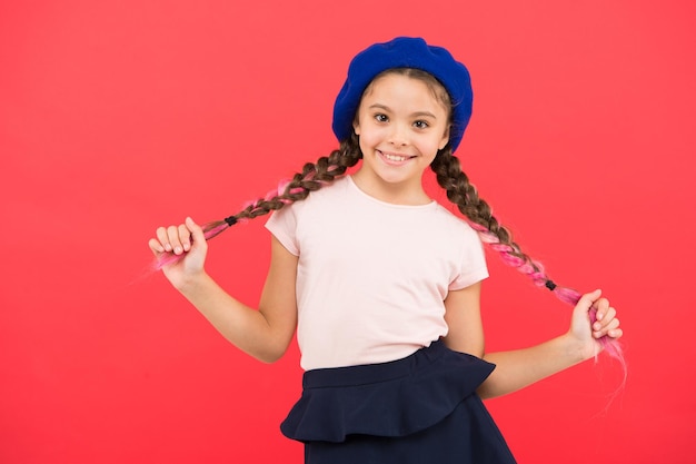 Kid little cute fashion girl posando con largas trenzas y sombrero de fondo rojo. Chica de moda. Accesorio de boina de moda. Moda adolescente. Atributo de la moda francesa. Niño niña pequeña feliz sonriente bebé.