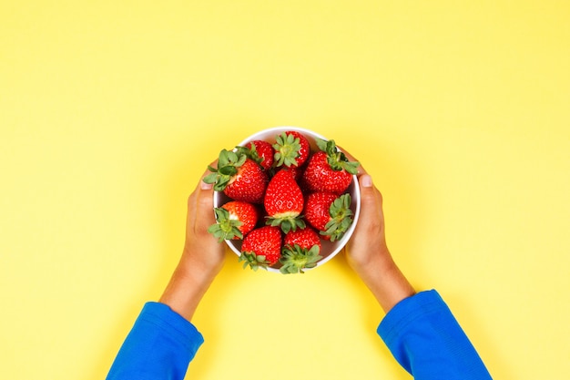 Kid sostiene en la mano tazón con fresas frescas sobre fondo amarillo