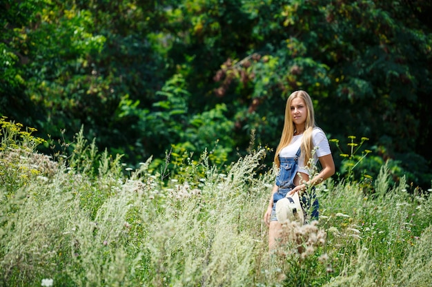 Kiev, Ucrania - Agosto de 2019 hermosa joven en overoles de mezclilla y un sombrero ligero caminando en el parque