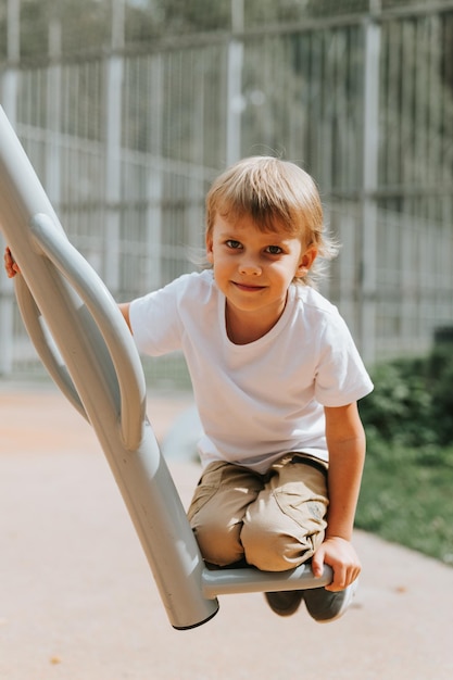 Kind auf dem Spielplatz der Stadt schaukelt auf einem Schaukelkarussell süßer kleiner glücklicher lächelnder offener fünfjähriger Junge mit langen blonden Haaren in einem weißen T-Shirt Generation z Kinder psychische Gesundheit Lebensstil