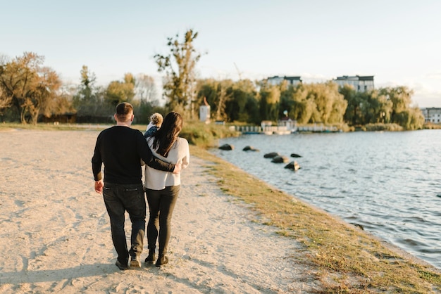 Kind auf den Händen der Eltern umarmt Vater zu Mutter Glücklicher Familienspaziergang am Strand bei Sonnenuntergang Kind, Tochter, Mutter, Vater, Spaziergang am Strand Familienfantasien Kindheitsträume Rückansicht