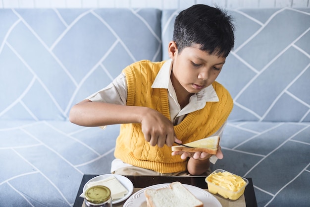 Kind bereitet sich das Frühstück vor, bevor es zur Schule geht. Fröhlicher Junge verteilt Butter auf Brot